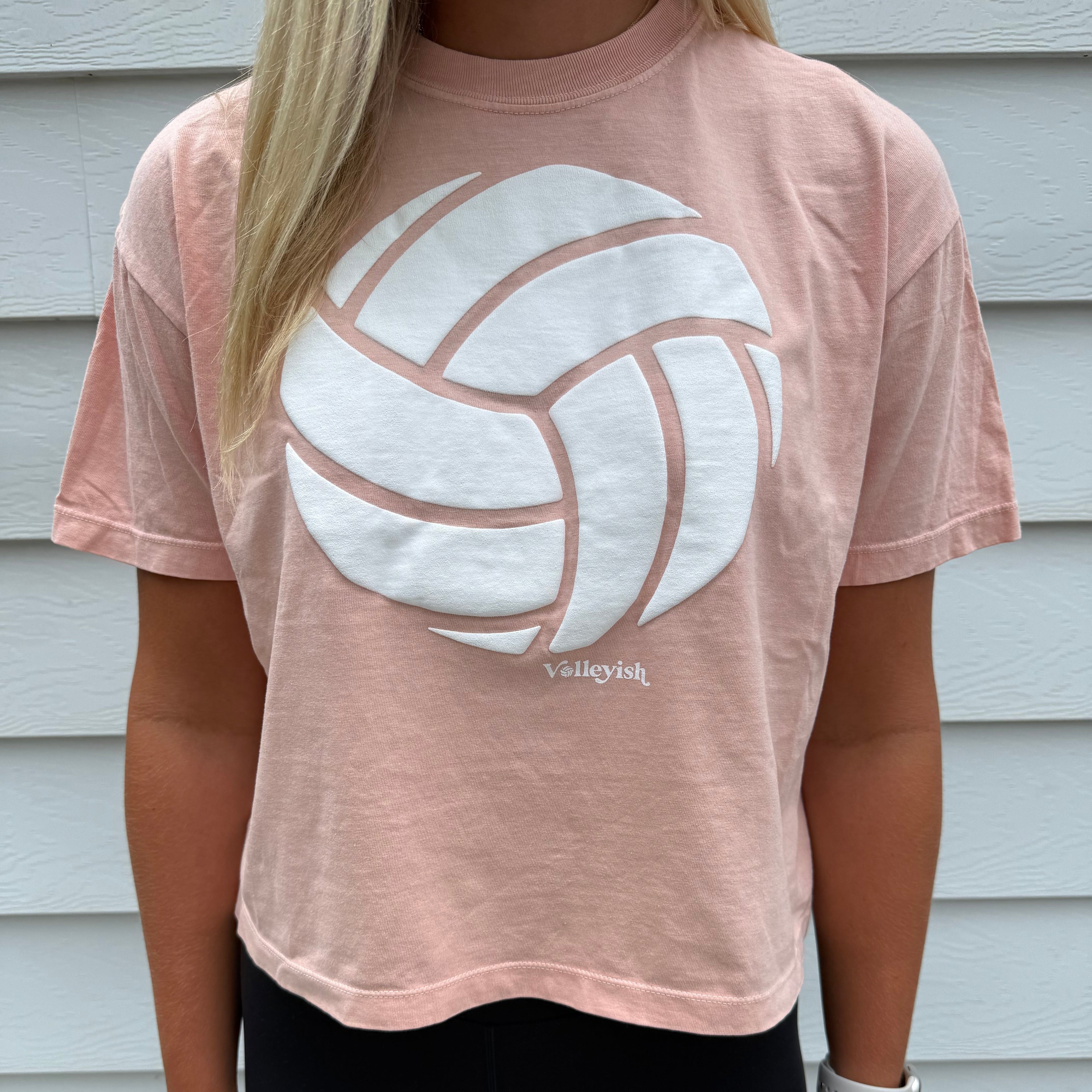 Woman wearing a peach t-shirt with a white volleyball graphic in front of a white wall