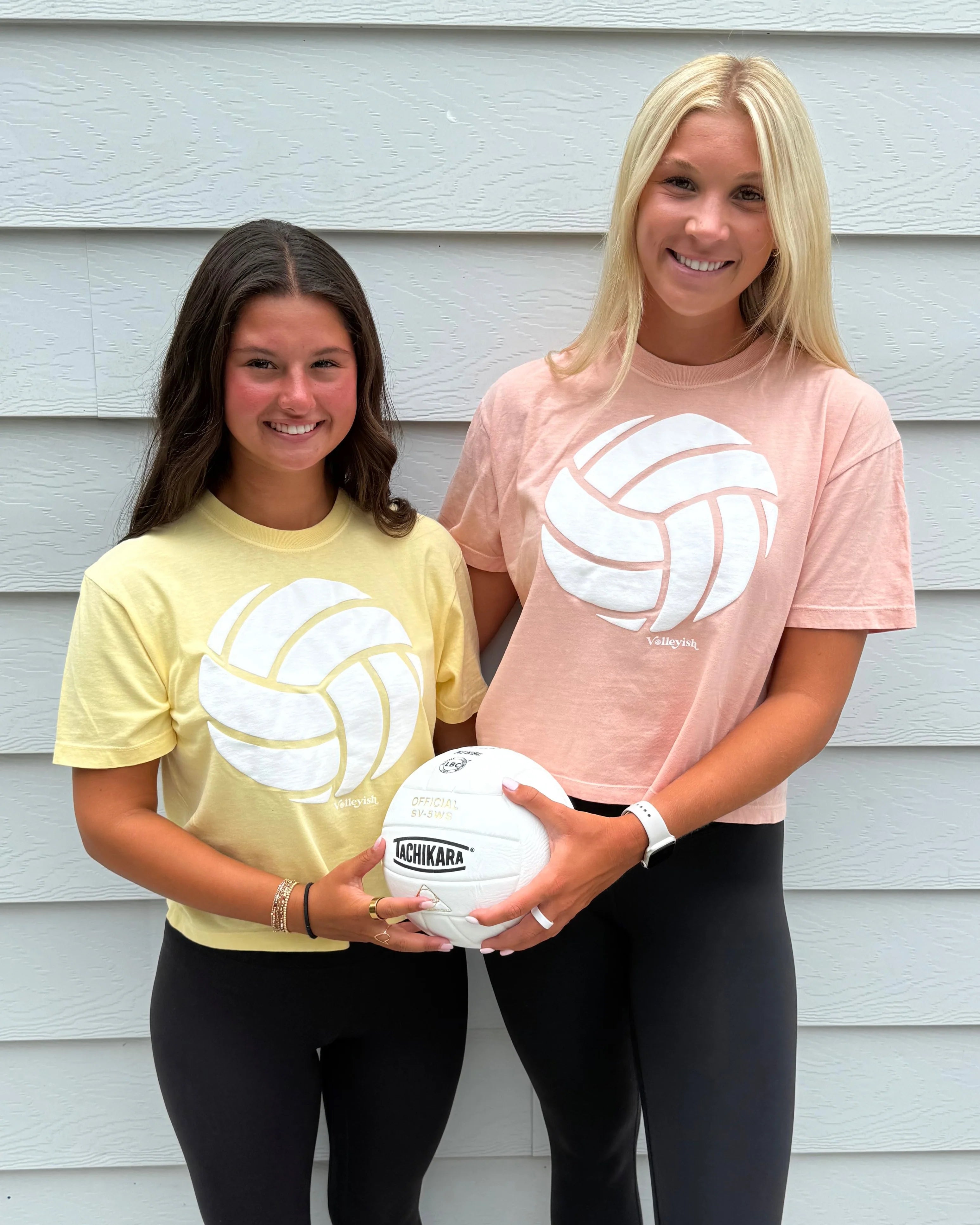 Two volleyball players wearing volleyball t-shirts and holding a volleyball and smiling