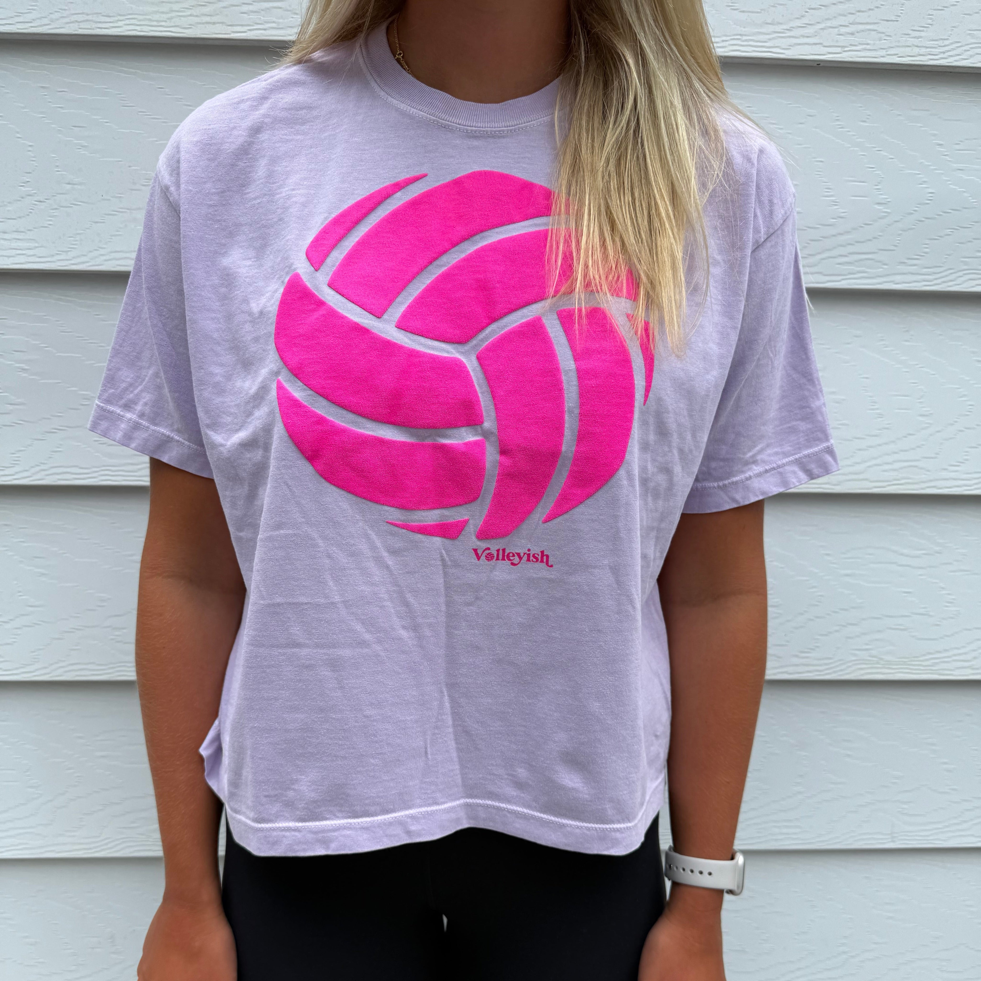 Person wearing a light purple t-shirt with a pink logo in front of a white wall.
