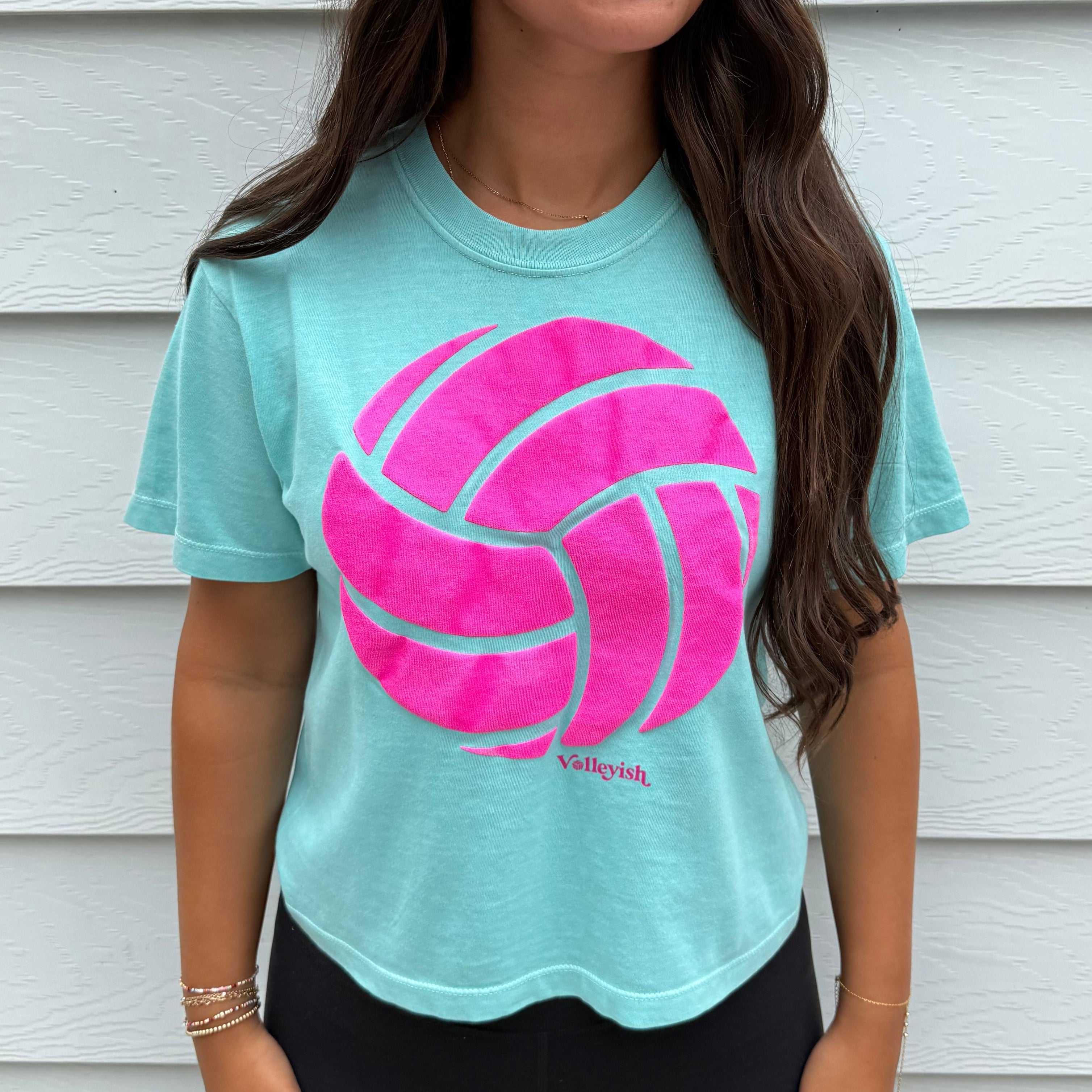 Woman wearing a turquoise t-shirt with a pink volleyball graphic in front of a white wall