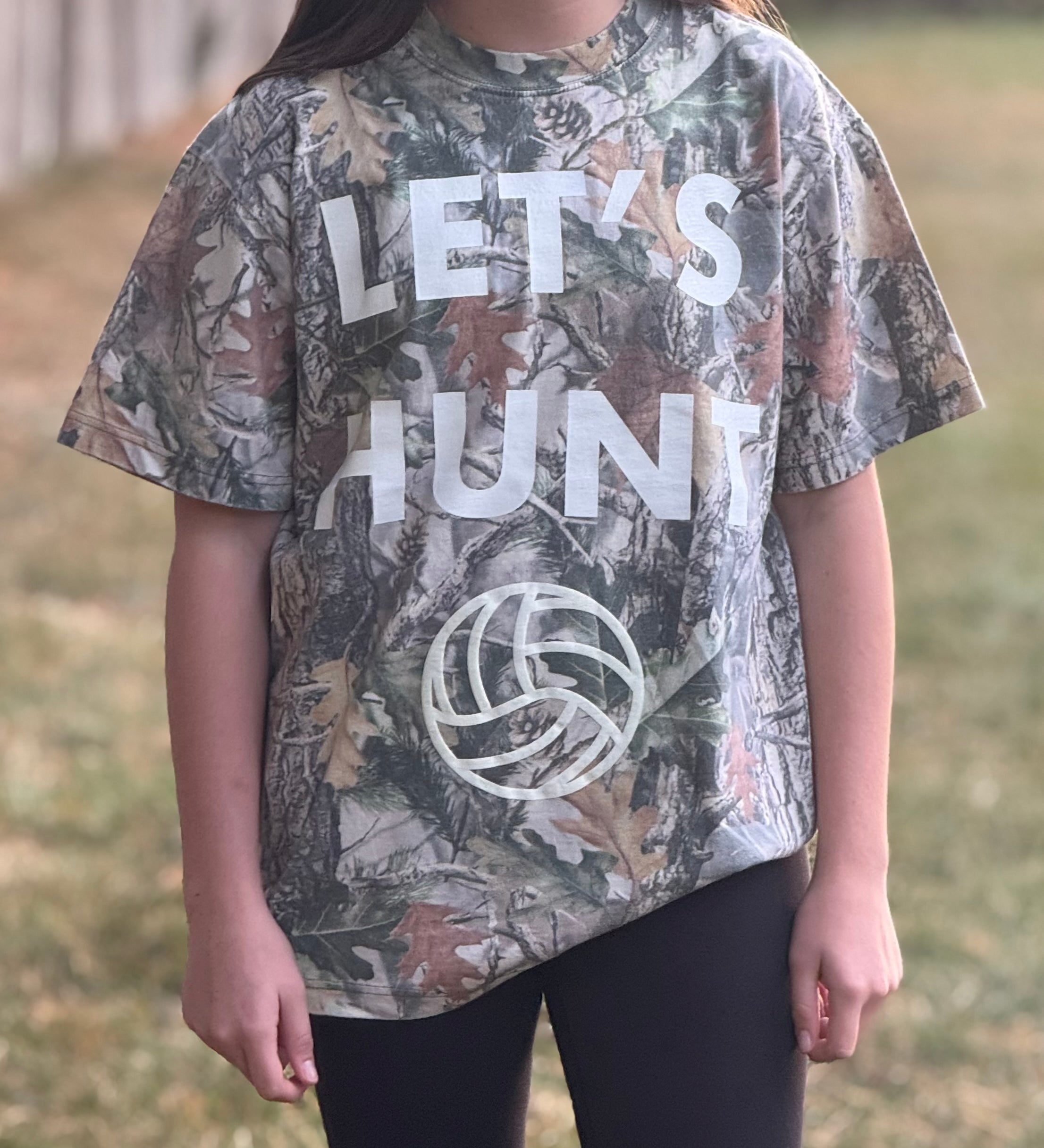 Volleyball girl wearing a camouflage t-shirt with 'Let's Hunt' text and volleyball graphic outdoors.
