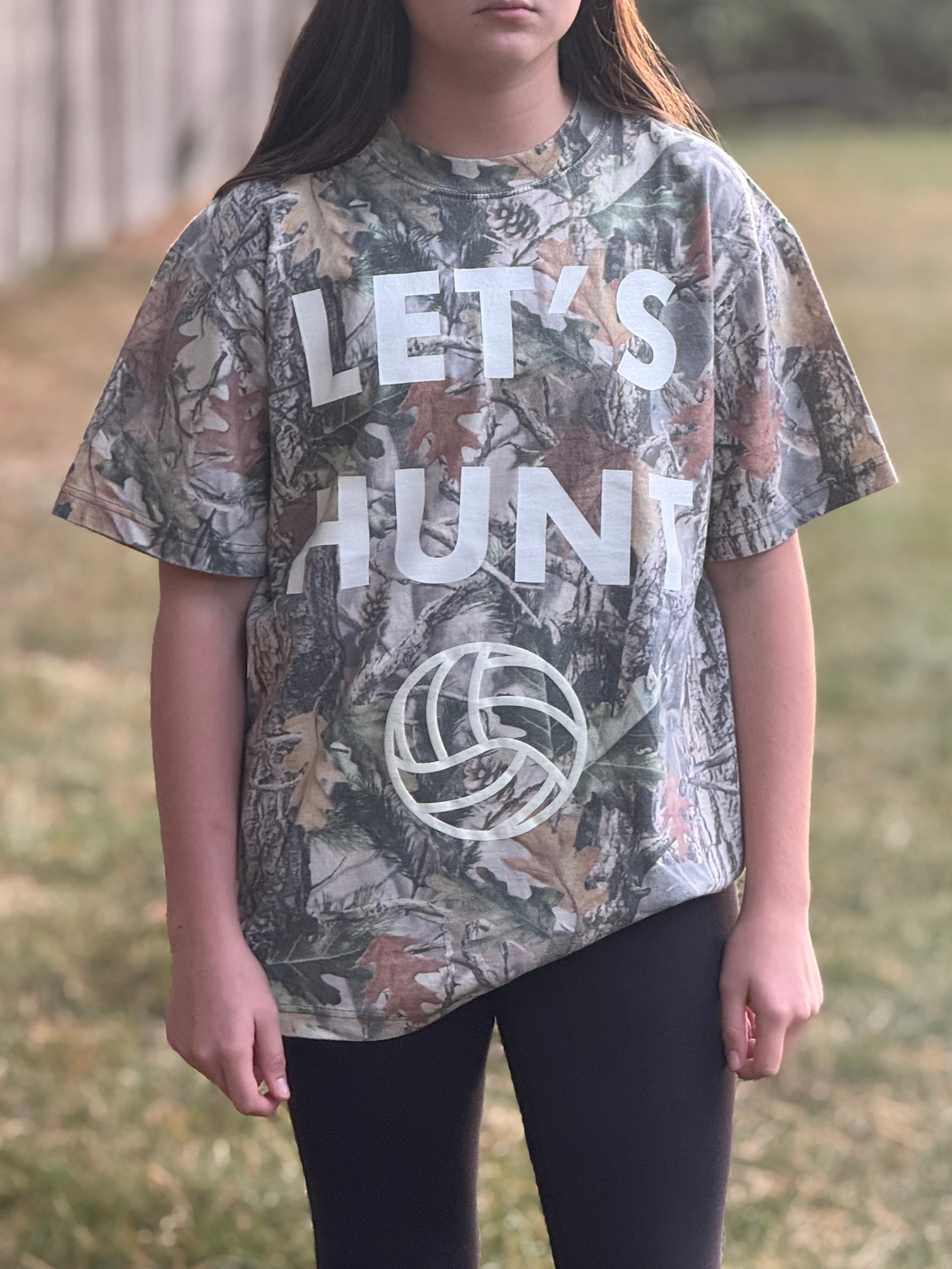 Volleyball girl wearing a camouflage t-shirt with 'Let's Hunt' text and volleyball graphic outdoors.