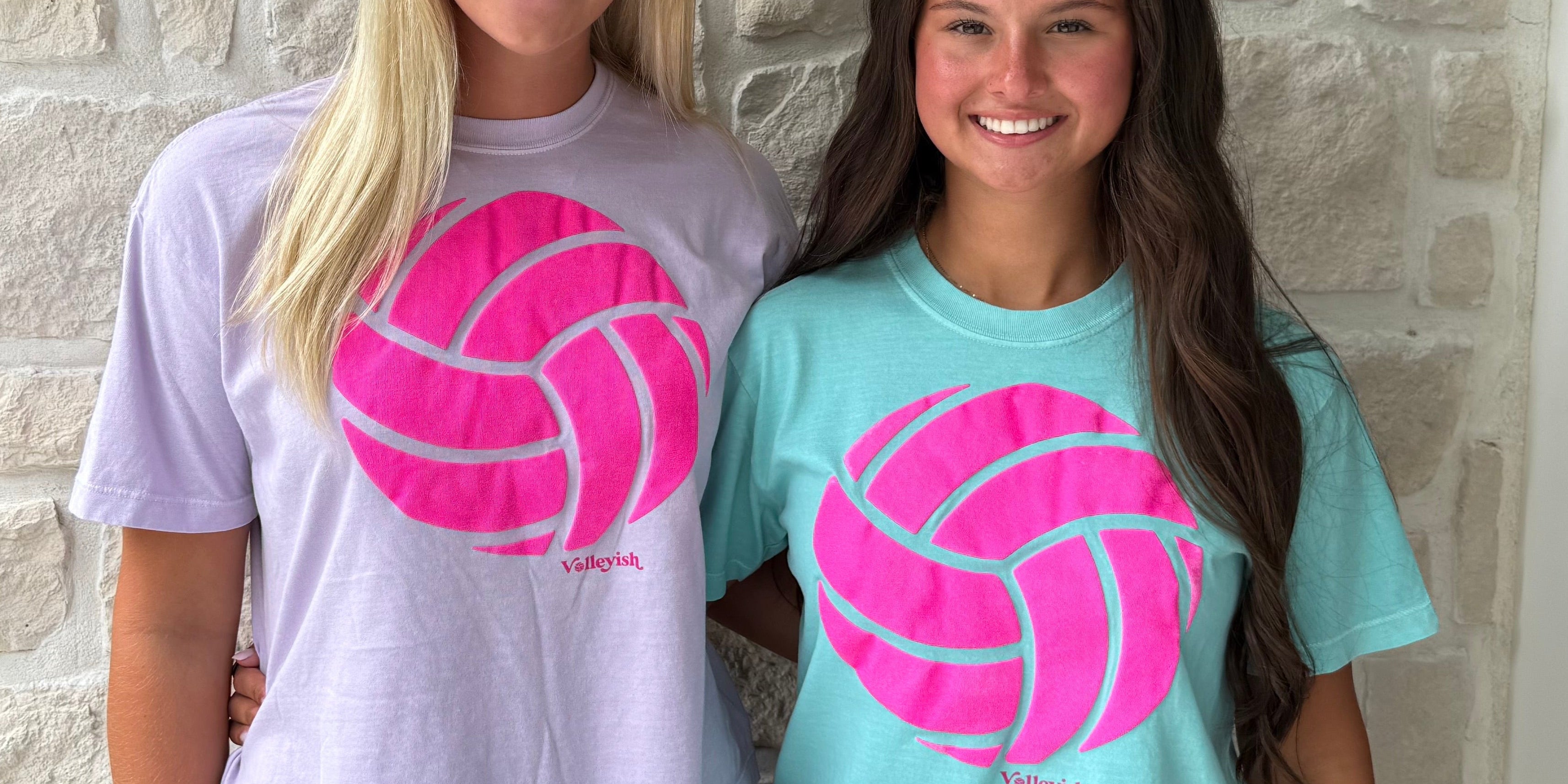 Two women wearing t-shirts with pink volleyball graphics standing in front of a stone wall.