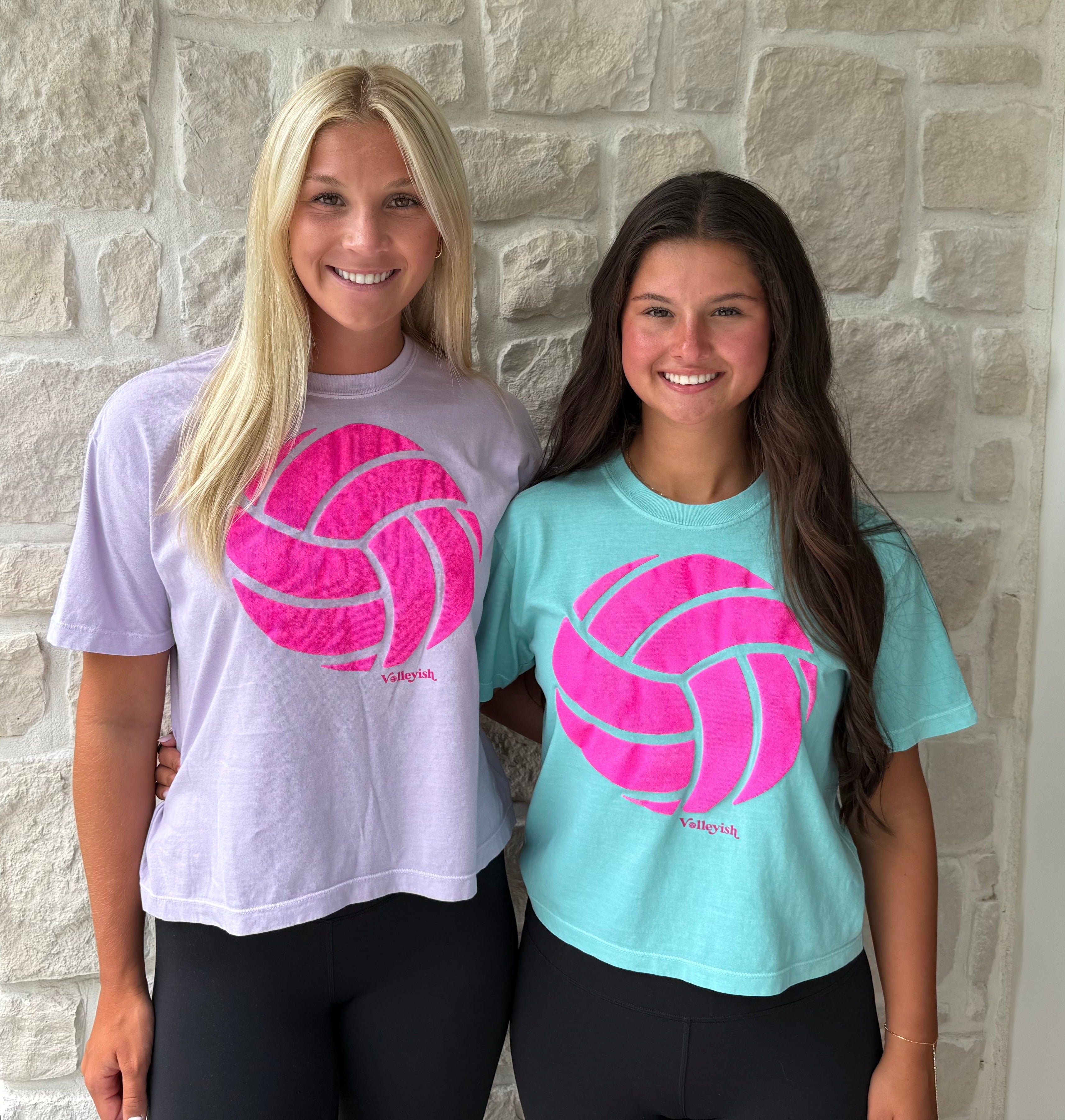 Two women wearing t-shirts with pink volleyball graphics standing in front of a stone wall.