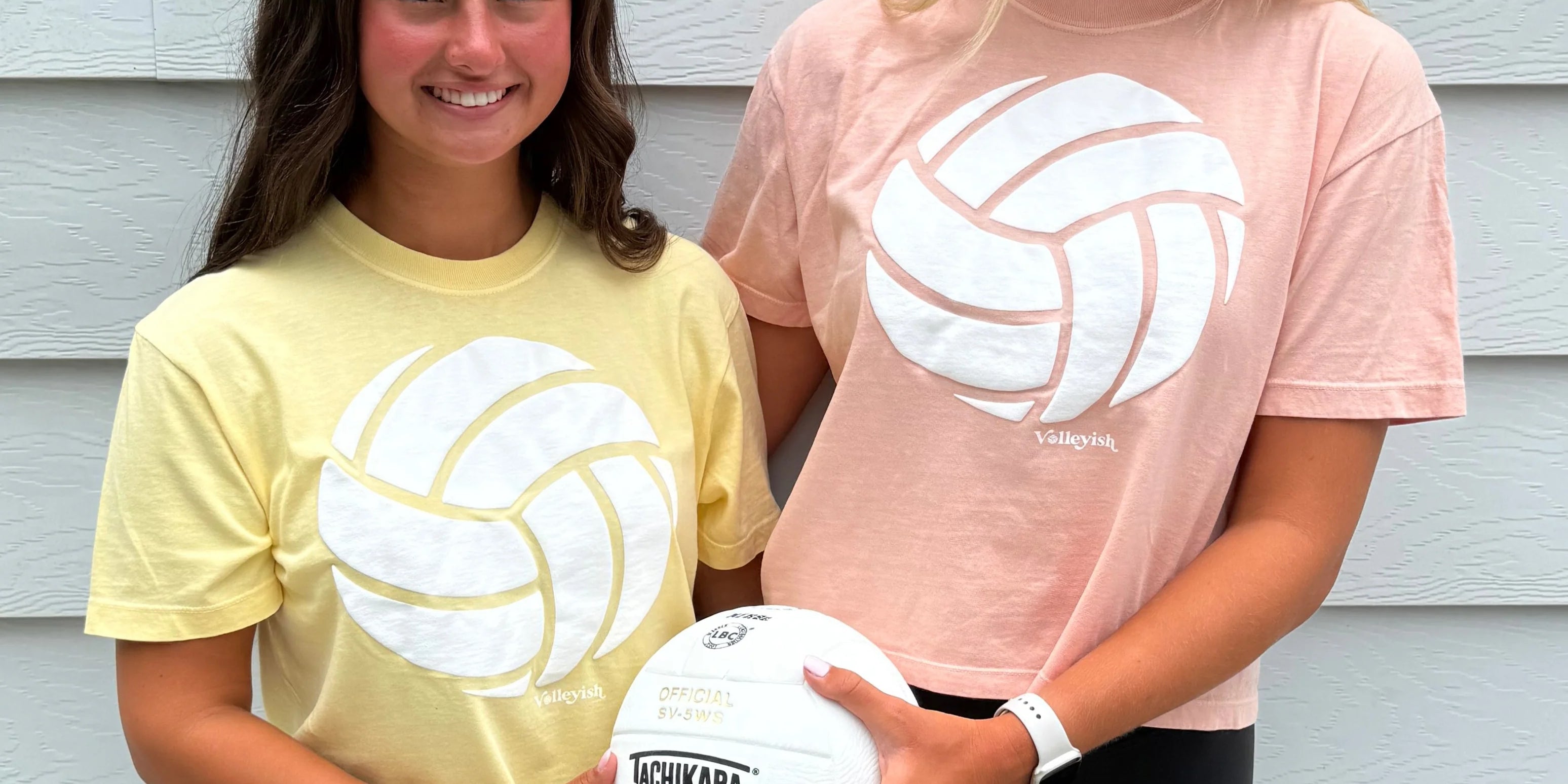 Two volleyball players wearing volleyball t-shirts and holding a volleyball and smiling
