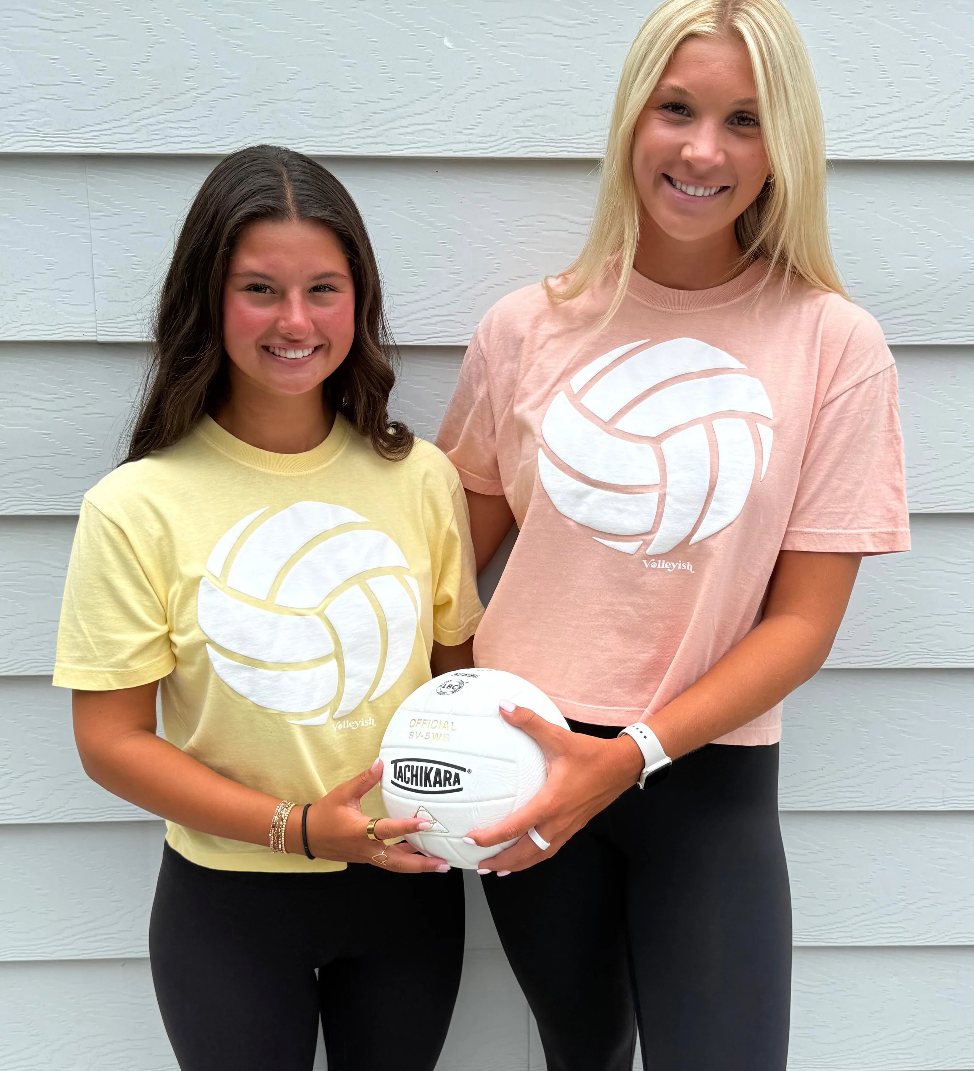 Two volleyball players wearing volleyball t-shirts and holding a volleyball and smiling